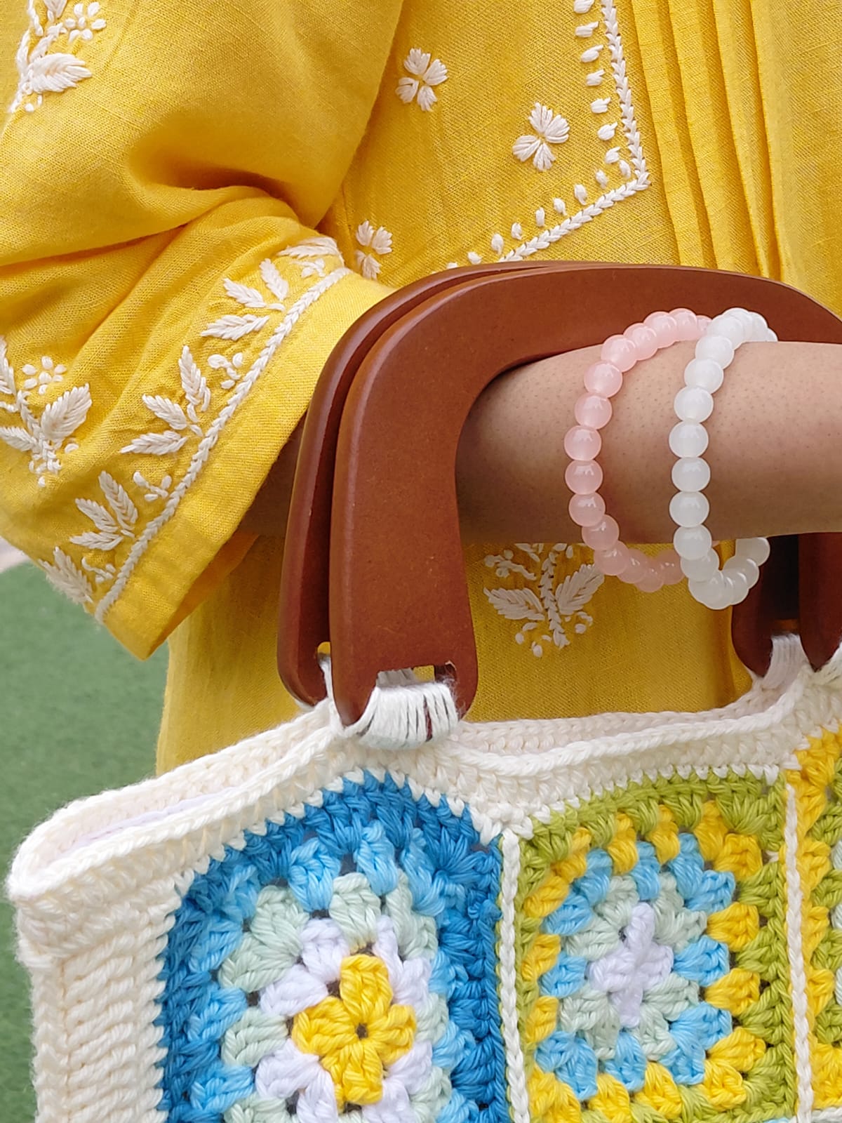 Handmade crochet granny square bag with a wooden handle. The tote features a multi-color floral pattern in blue, yellow, and white, with a cream-colored border. A woman is holding the boho-style bag with a yellow kurta.