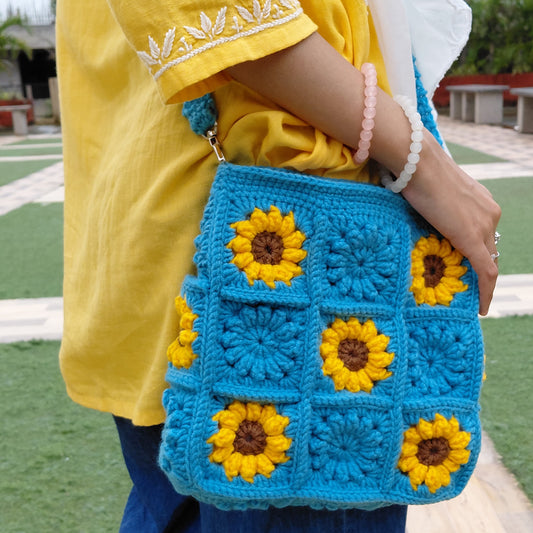 Handmade crochet sunflower bag in bright blue. The purse features yellow and brown sunflower granny squares, a long shoulder strap, and a square shape. A woman is wearing the bag outdoors.

