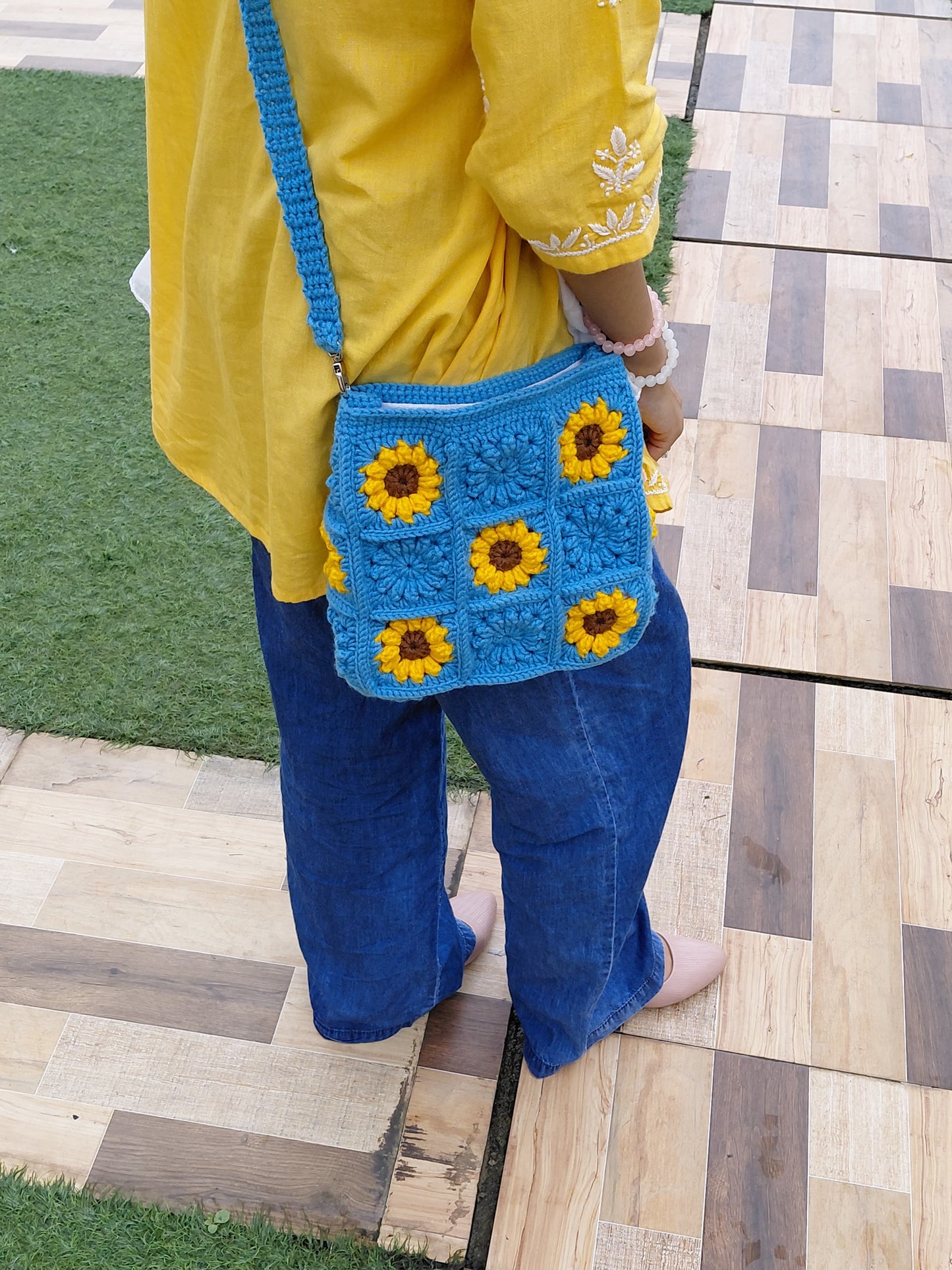 Handmade crochet sunflower bag in bright blue. The purse features yellow and brown sunflower granny squares, a long shoulder strap, and a square shape. A woman is wearing the bag outdoors.

