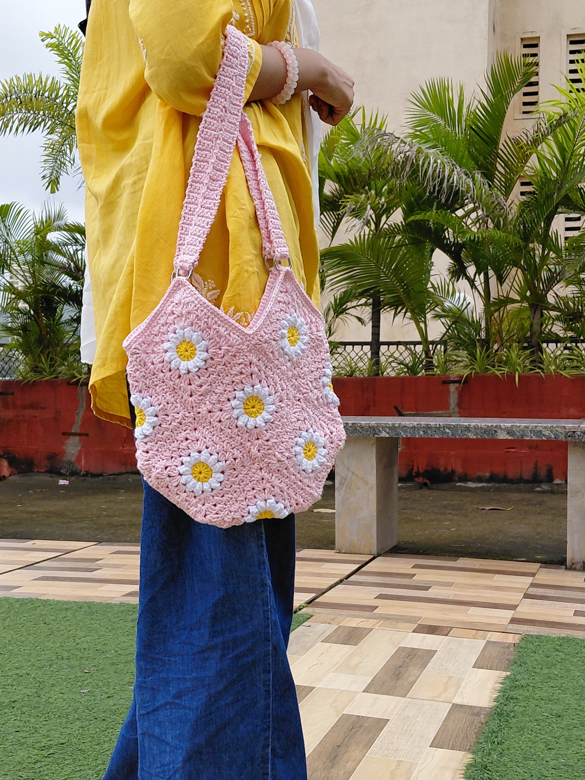 Handmade crochet daisy granny square bag in light pink. A woman is wearing the floral bag, which features white and yellow daisy motifs, a double strap, and a zipper closure. It is a perfect summer tote or boho purse accessory.

