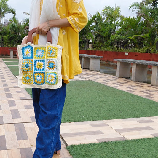 Handmade crochet granny square bag with a wooden handle. The tote features a multi-color floral pattern in blue, yellow, and white, with a cream-colored border. A woman is holding the boho-style bag with a yellow kurta.