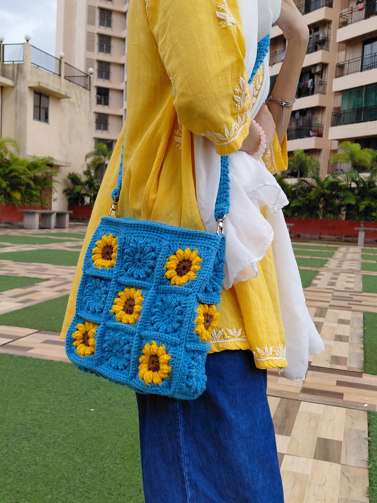 Handmade crochet sunflower bag in bright blue. The purse features yellow and brown sunflower granny squares, a long shoulder strap, and a square shape. A woman is wearing the bag outdoors.

