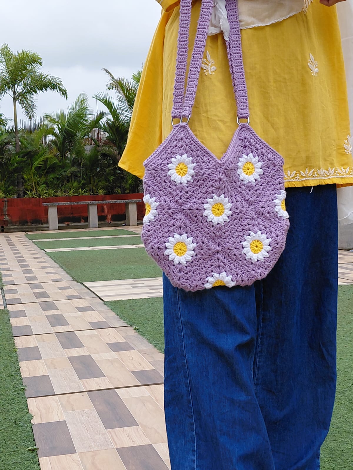 Handmade crochet daisy granny square bag in lavender purple. A woman is wearing the floral bag, which features white and yellow daisy motifs, a double strap, and a zipper closure. It is a perfect summer tote or boho purse accessory.