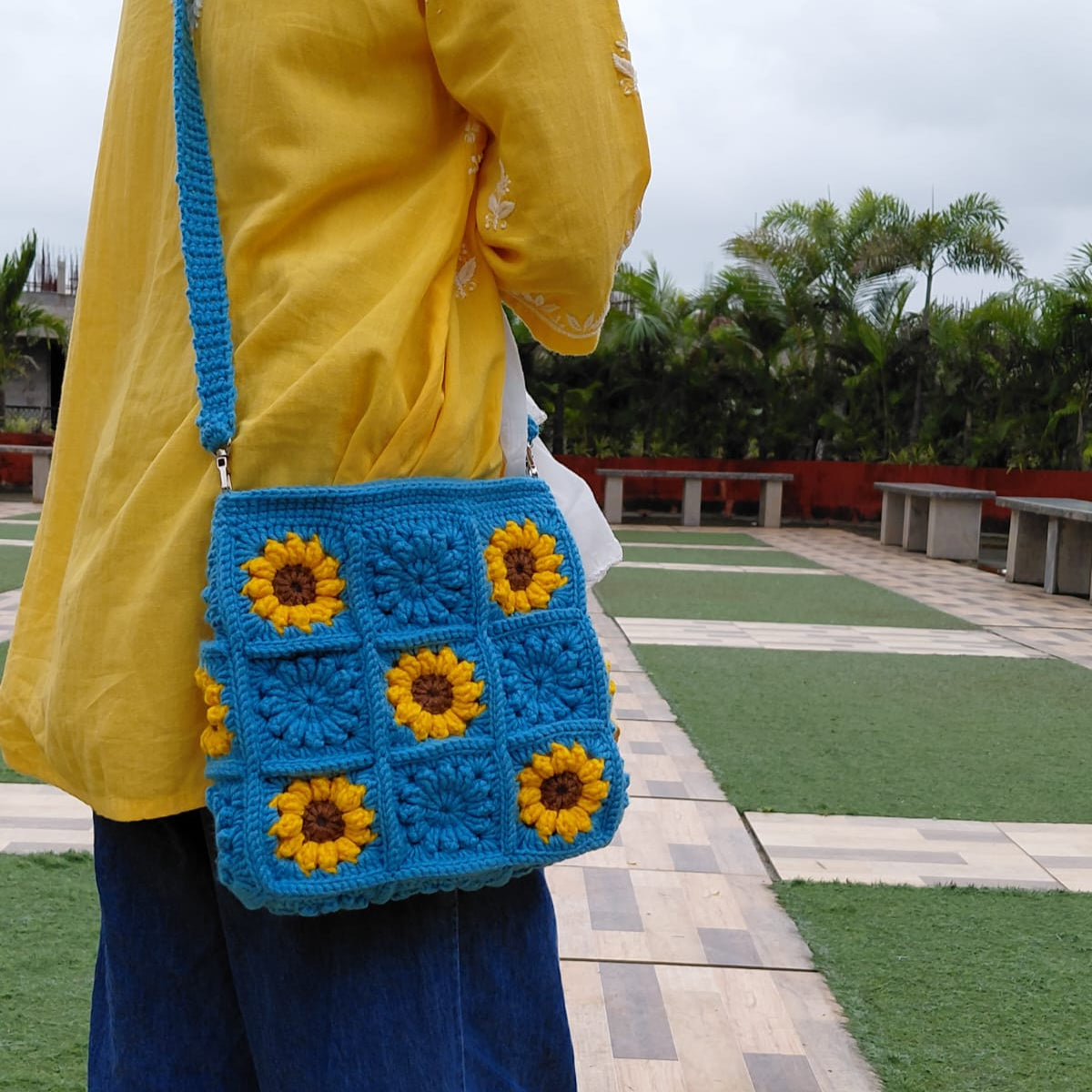 Handmade crochet sunflower bag in bright blue. The purse features yellow and brown sunflower granny squares, a long shoulder strap, and a square shape. A woman is wearing the bag outdoors.

