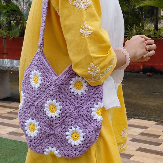 Handmade crochet daisy granny square bag in lavender purple. A woman is wearing the floral bag, which features white and yellow daisy motifs, a double strap, and a zipper closure. It is a perfect summer tote or boho purse accessory.