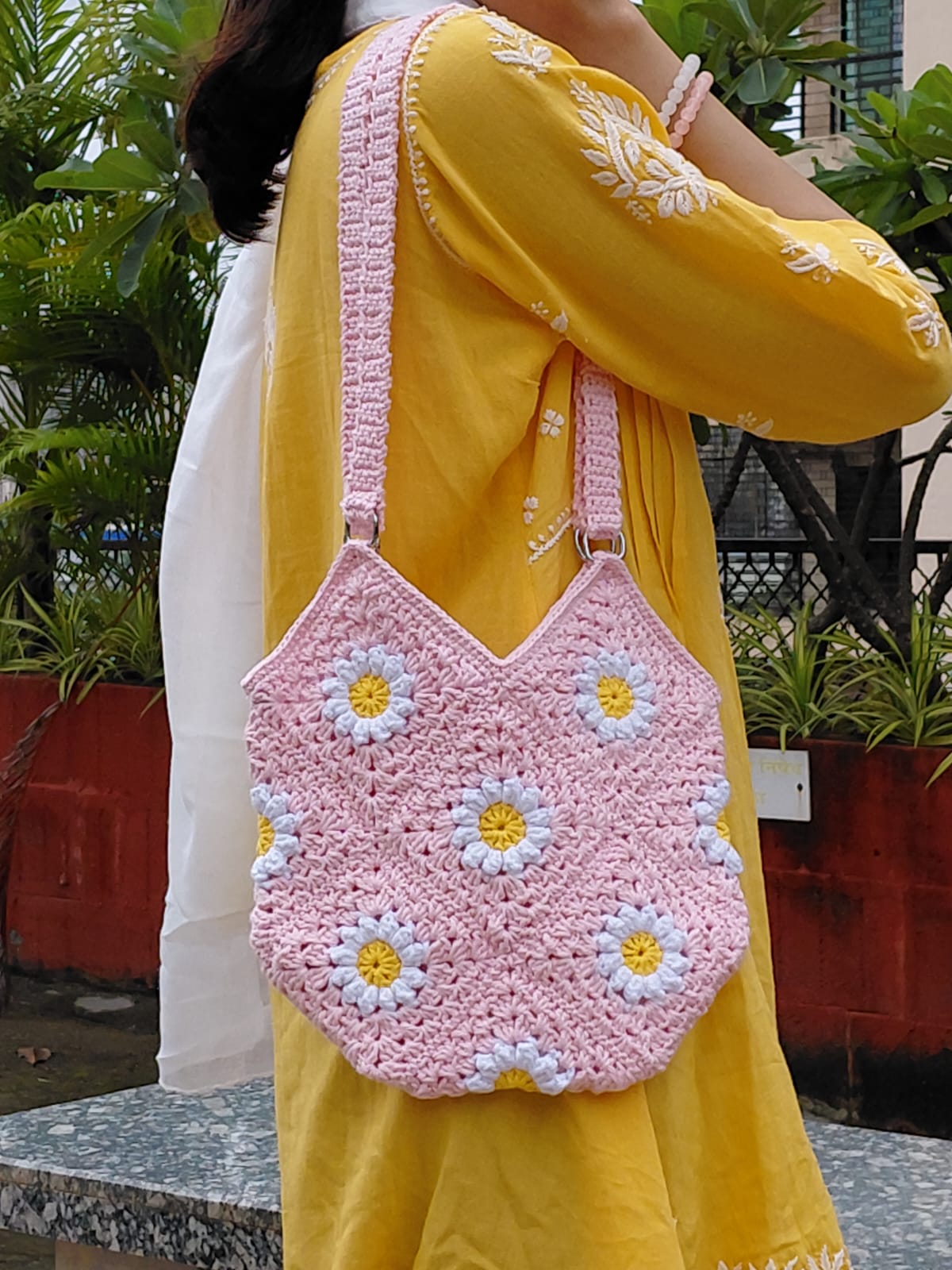Handmade crochet daisy granny square bag in light pink. A woman is wearing the floral bag, which features white and yellow daisy motifs, a double strap, and a zipper closure. It is a perfect summer tote or boho purse accessory.

