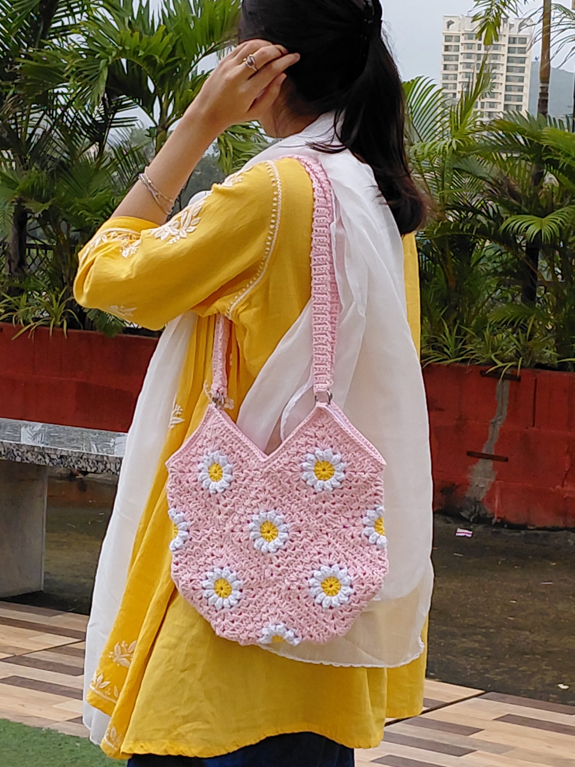 Handmade crochet daisy granny square bag in light pink. A woman is wearing the floral bag, which features white and yellow daisy motifs, a double strap, and a zipper closure. It is a perfect summer tote or boho purse accessory.
