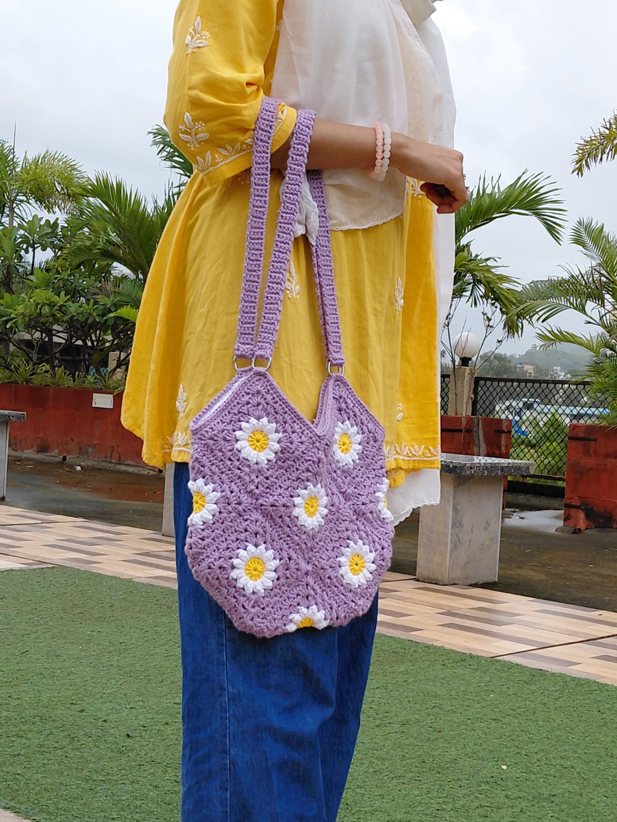 Handmade crochet daisy granny square bag in lavender purple. A woman is wearing the floral bag, which features white and yellow daisy motifs, a double strap, and a zipper closure. It is a perfect summer tote or boho purse accessory.