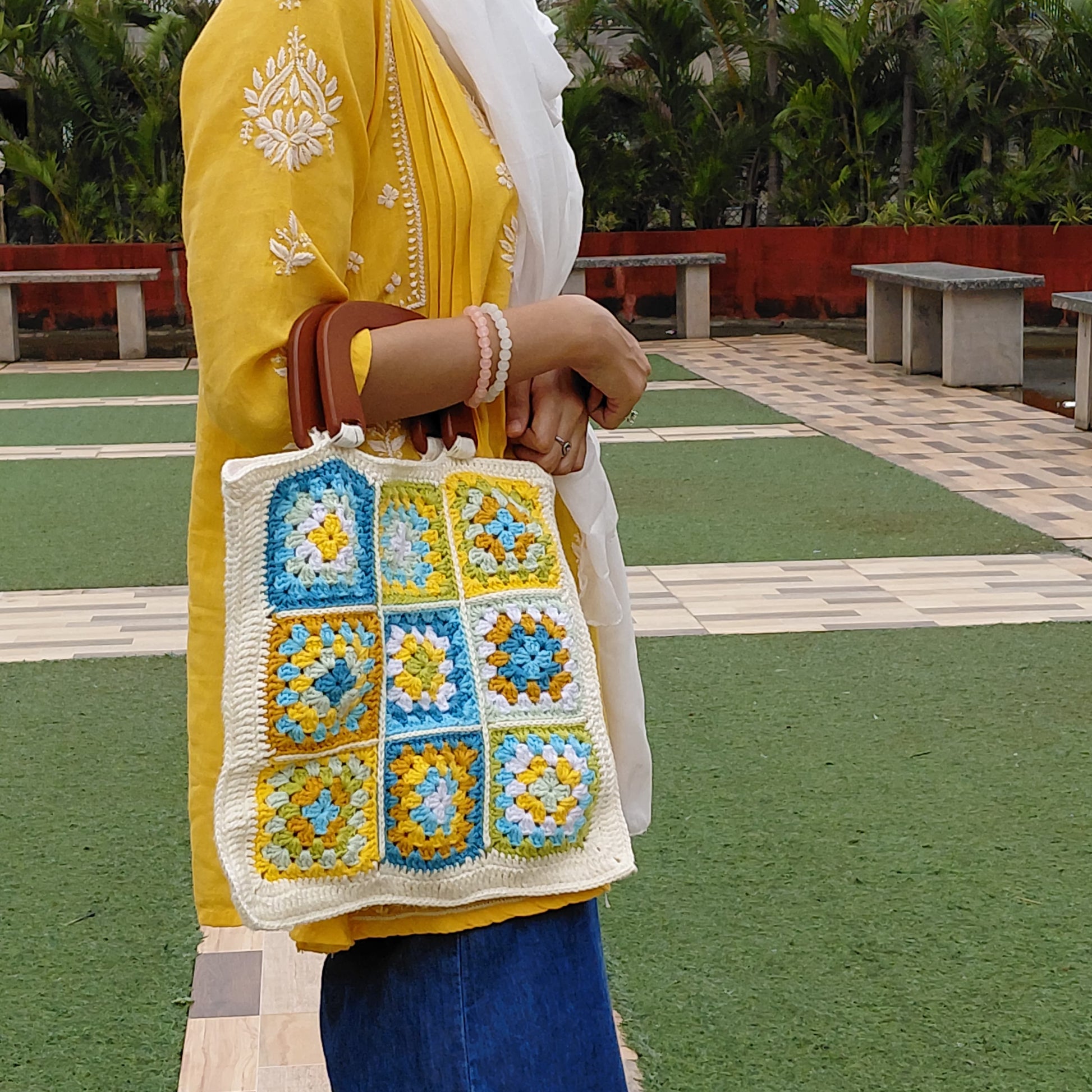 Handmade crochet granny square bag with a wooden handle. The tote features a multi-color floral pattern in blue, yellow, and white, with a cream-colored border. A woman is holding the boho-style bag with a yellow kurta.